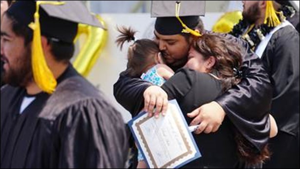 A man in a cap and gown at his graduation ceremony hugging his wife and child.