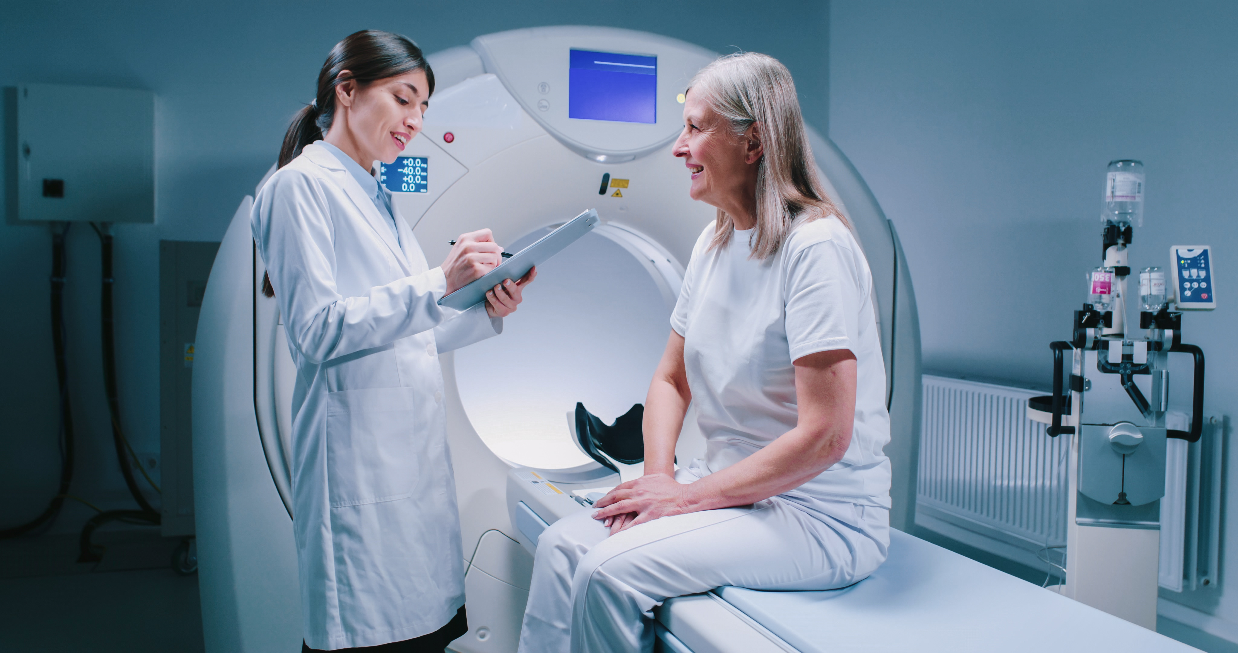 Two women talking, one a patient sitting on an MRI machine and the other a doctor with a clipboard.